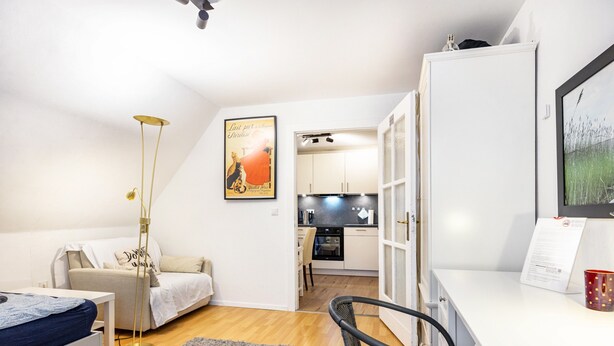 Ein gemütlicher Raum mit Bett, Schreibtisch, Stuhl, Stehlampe und einer Küchenzeile im Hintergrund.