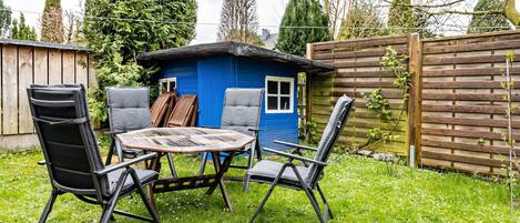 A blue wooden structure with a window, surrounded by a wooden fence, and a table with chairs in a grassy yard.