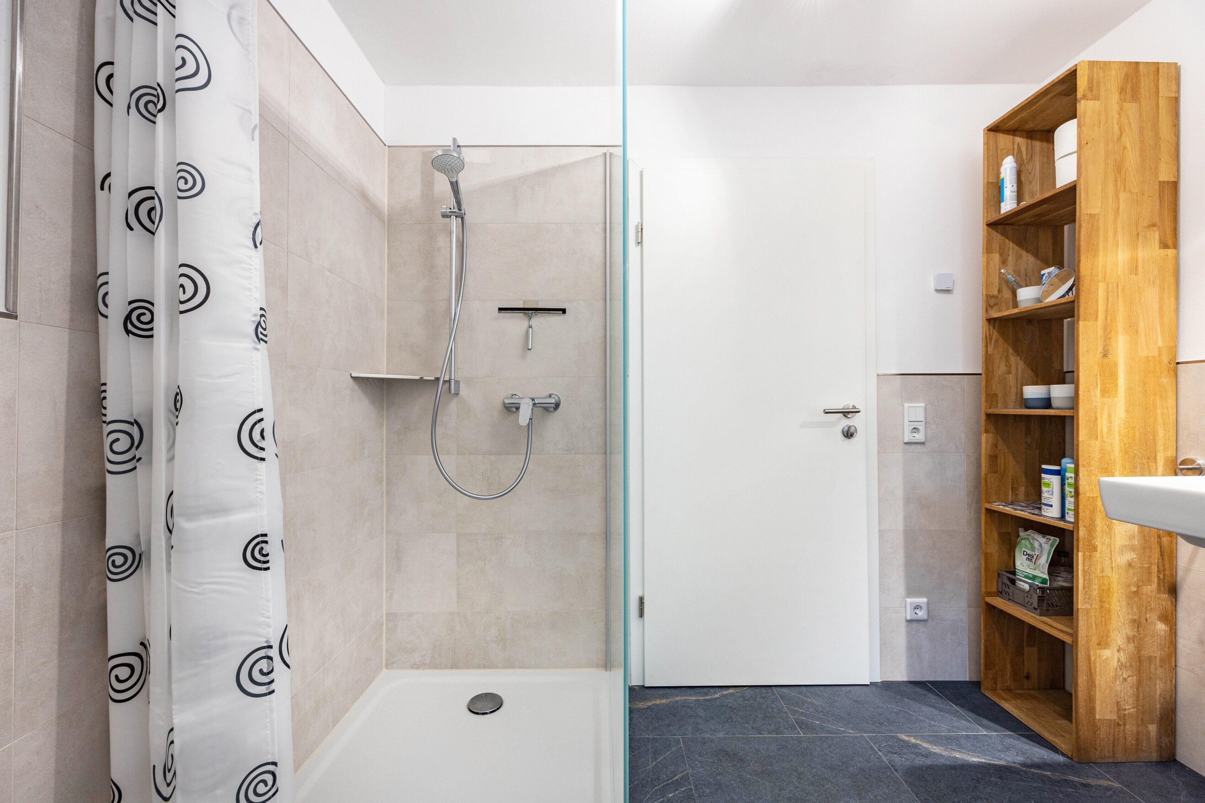 A bathroom with a shower, a bathtub, a wooden shelf with toiletries, and a white sink.