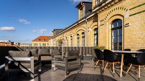 Un patio exterior con un sofá, sillas y una mesa, con vistas a un edificio histórico de fachada de ladrillo y ventanas arqueadas.
