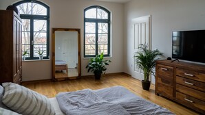 Un dormitorio con cama, cómoda, televisor, plantas en macetas y dos ventanas de arco.