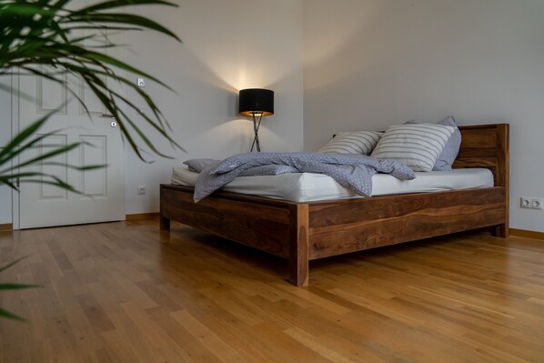 Un lit en bois avec une tête de lit, une lampe de chevet et une plante dans une pièce dont le sol est recouvert de planches de bois et qui possède une porte blanche.