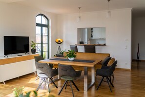 A modern living room with a TV, dining table, chairs, a plant, and a large window. - Apartment 'König Albert Residenz' with Private Terrace, Balcony and Wi-Fi (Leipzig)