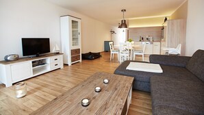 Ein modernes Wohnzimmer mit einem Flachbildfernseher, einem Holztisch mit Kerzen, einem grauen Sofa und einem Essbereich mit weißen Stühlen.