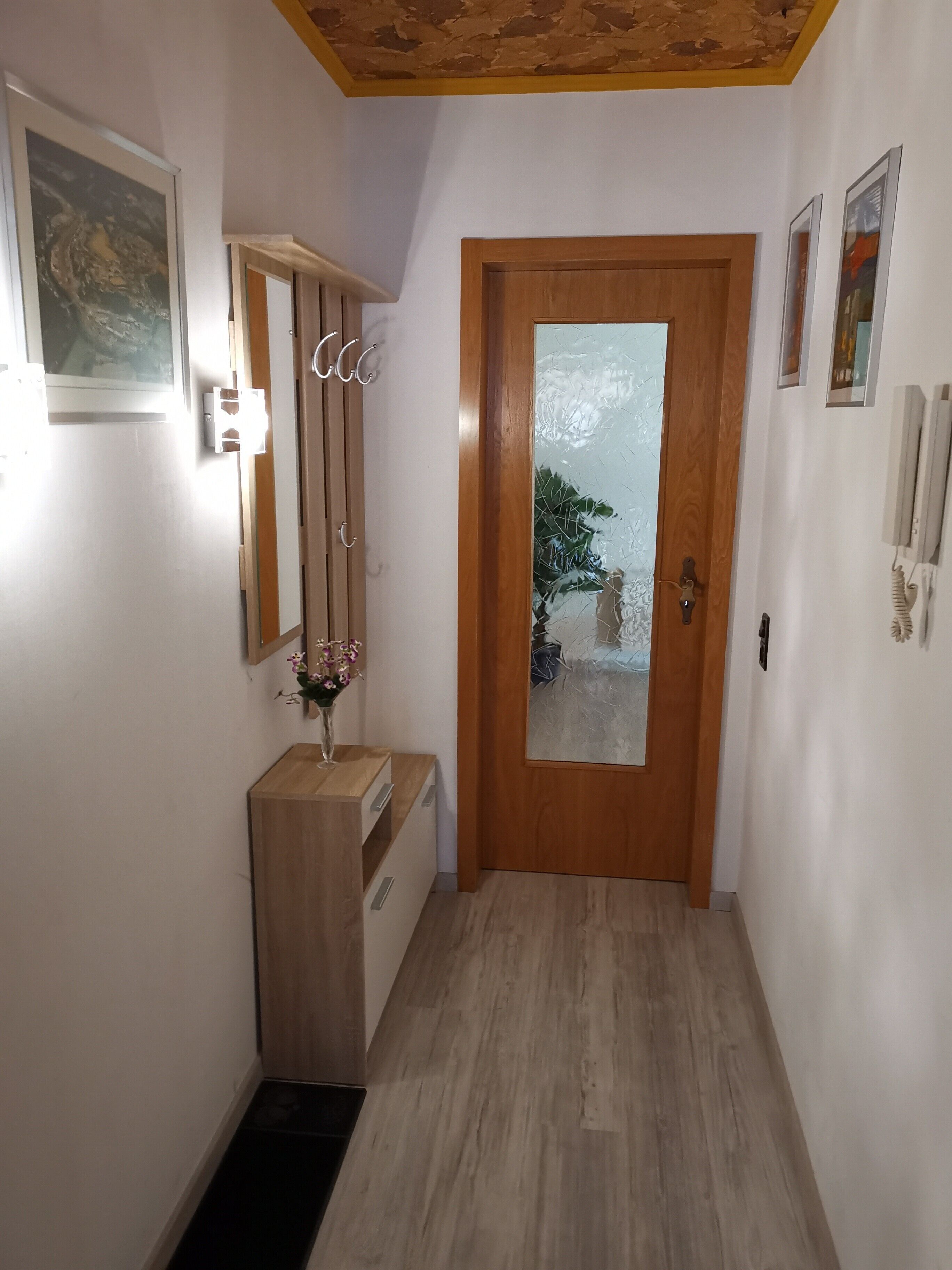 A hallway with a wooden door, a small wooden desk, a vase with flowers, and a framed picture on the wall.