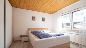 Una camera da letto con soffitto in legno, un letto con materasso bianco e biancheria azzurra, due quadri appesi al muro e una finestra vista edifici.