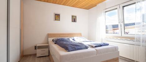 A bedroom with a wooden ceiling, a bed with a white mattress and blue bedding, two framed pictures on the wall, and a window with a view of buildings.