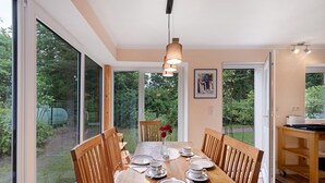 A dining room with a large table set for six, wooden chairs, a hanging light fixture, and a view of a garden through large windows.