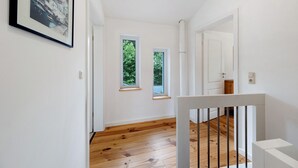 A hallway with wooden flooring, white walls, and two windows with views of greenery.