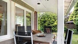 A covered patio with a wooden table and chairs, a glass door, and a view of a garden.