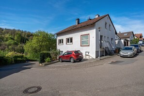 Une rue résidentielle avec une maison blanche, une voiture rouge garée devant, et une autre voiture garée sur le côté.