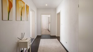 Ein Flur mit weißer Wand, dunklem Boden und einer Tür am Ende. Drei Gemälde hängen an der Wand, und auf einem kleinen Tisch steht ein weißes Objekt.