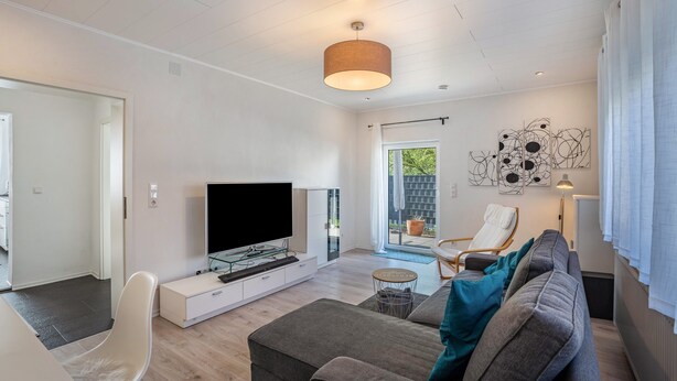 Ein modernes Wohnzimmer mit Flachbildfernseher, einer Couch, einem Esstisch und einem Sessel.
