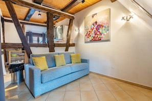 Una habitación con sofá azul, vigas de madera, suelo de baldosas, un cuadro y una lámpara fijada a la pared.
