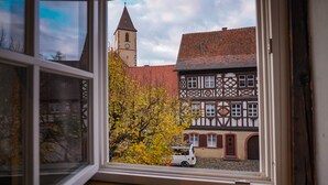 Vista dalla finestra di una casa tradizionale a graticcio, con una torre dell'orologio, un albero con foglie gialle e un furgone bianco parcheggiato davanti.