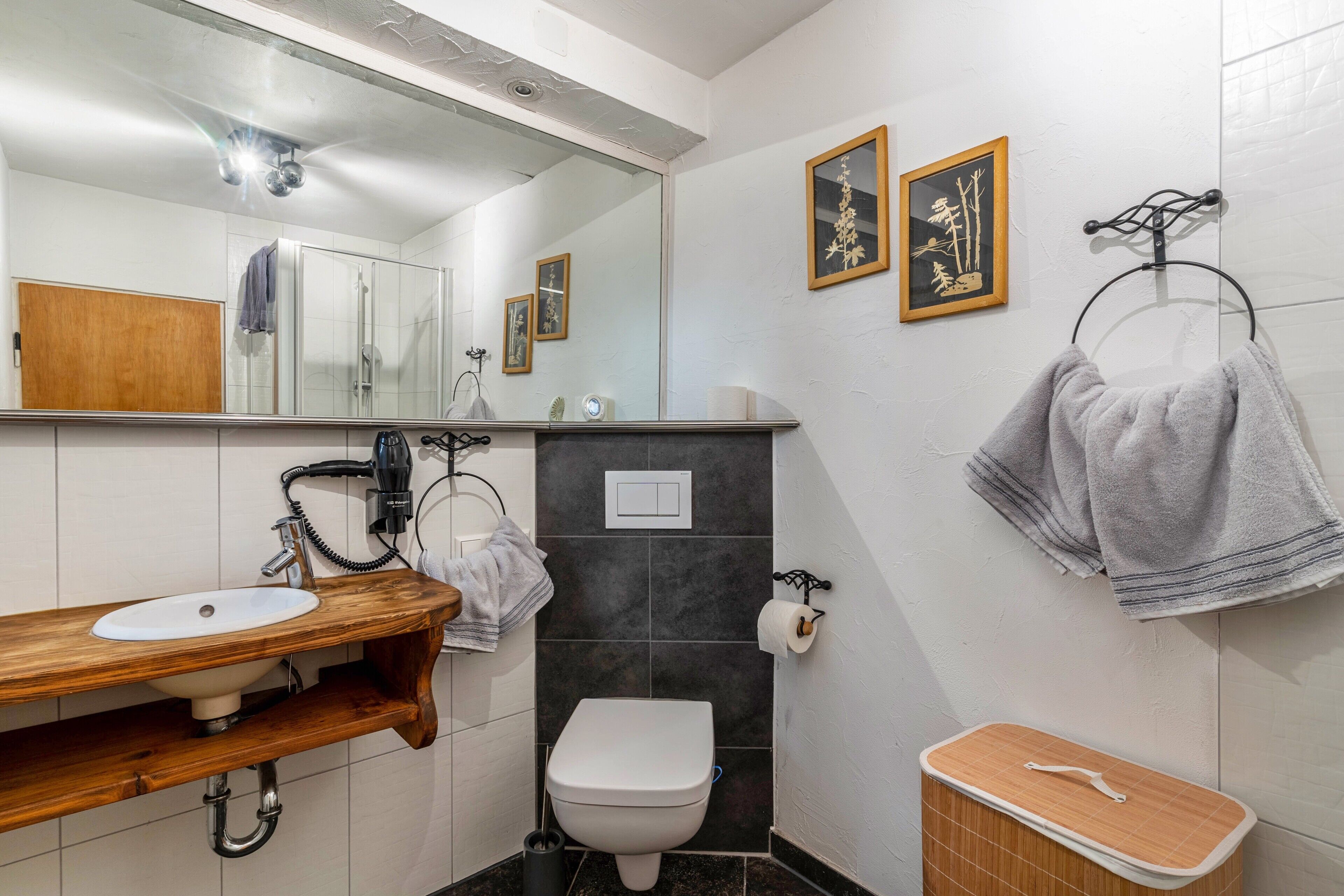 Un baño con tocador de madera, un lavabo blanco, un inodoro y un espejo. Hay toallas colgadas en un perchero y un cuadro en la pared.