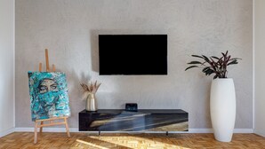 Ein Zimmer mit Holzboden, einem Gemälde auf einem Staffelei, einem Flachbildfernseher an der Wand, einem Konsolentisch und einer großen Vase mit Pflanzen.