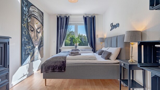 Ein Schlafzimmer mit Bett, Nachttisch, Lampe, einem Fenster mit Vorhängen und einem Wandbild.