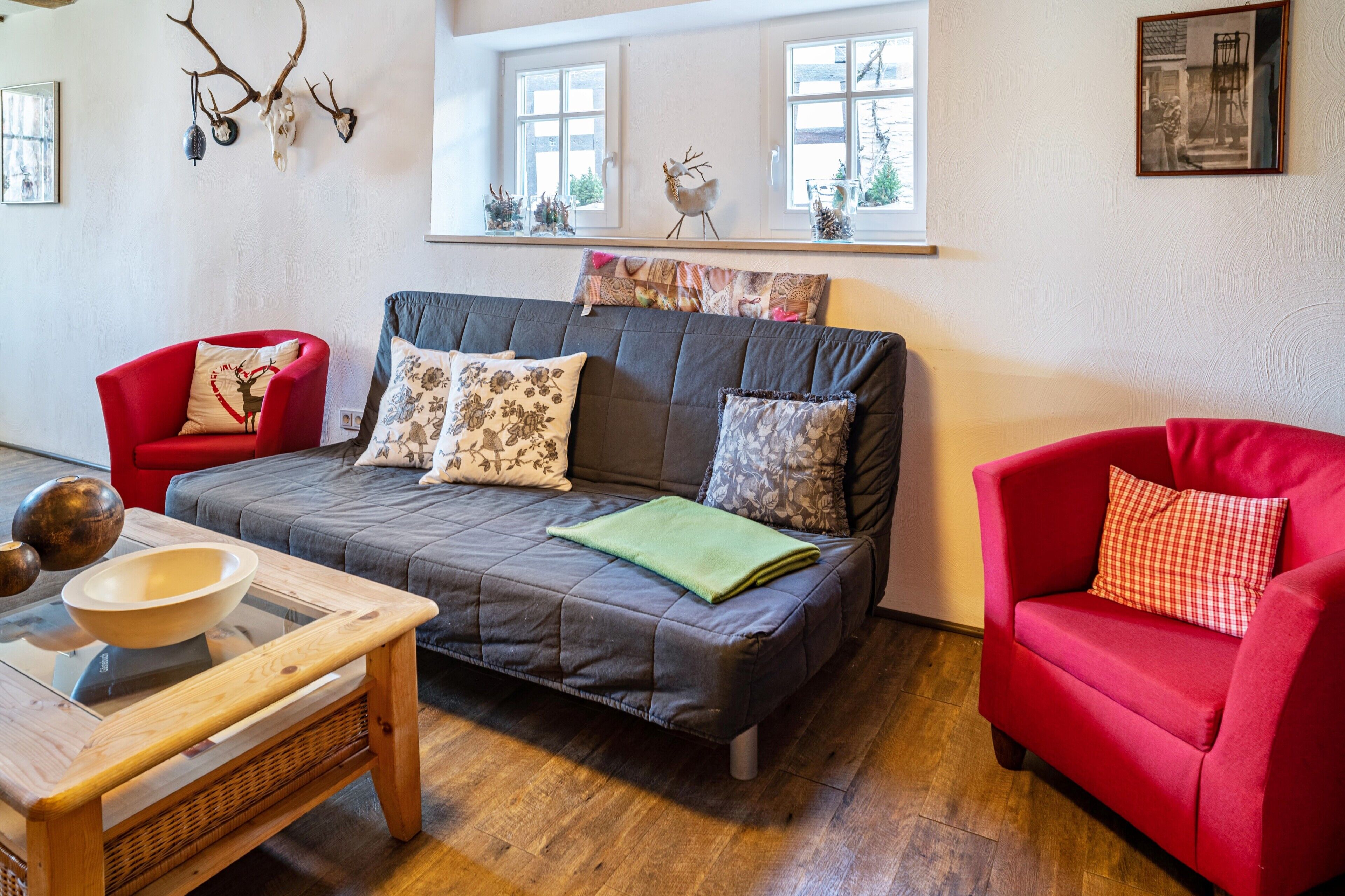 Ein gemütliches Wohnzimmer mit grauem Sofa, roten Sesseln, einem Holztisch und einem Fenster mit Dekoration.