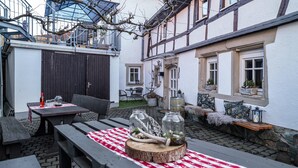 An outdoor patio with wooden benches, a table with a red and white checkered tablecloth, and potted plants. - Vacation Home "Gästehaus Vis À Vis" with Mountain View, Shared Garden & Wi-Fi (Wallhausen)