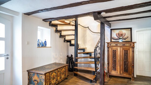 A staircase with wooden steps and a metal railing, a wooden cabinet with a vase, and a decorative chest in a room with wooden flooring and white walls. - Vacation Home "Gästehaus Vis À Vis" with Mountain View, Shared Garden & Wi-Fi (Wallhausen)