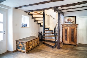 Un escalier avec des marches en bois et une rampe en métal, une armoire en bois ornée d’un vase, et une commode décorative dans une pièce au plancher en bois et aux murs blancs.