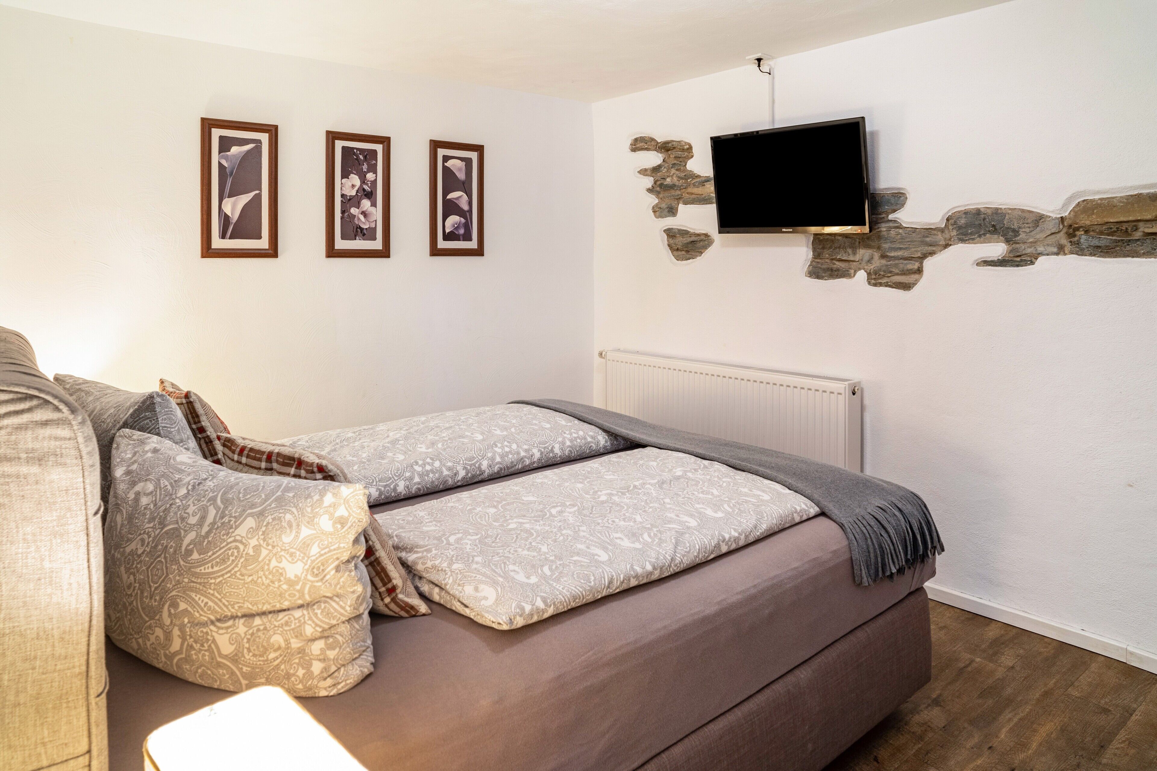 A bedroom with a bed, a television mounted on a stone wall, and three framed pictures on the wall.