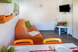 A room with a sofa, a table with chairs, a television mounted on the wall, and a clock.