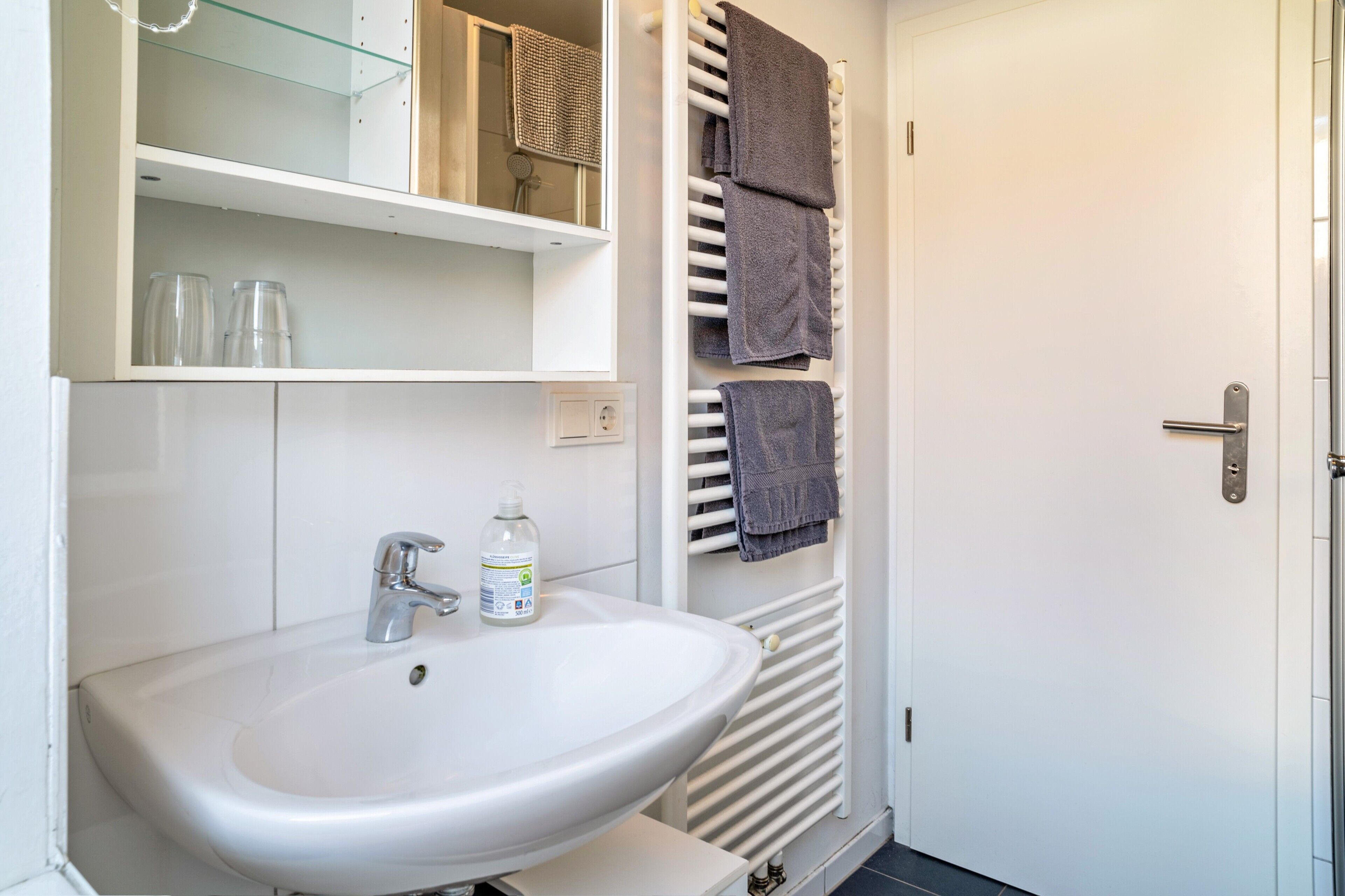A bathroom with a white sink, a glass shelf, a towel rack, and a door.