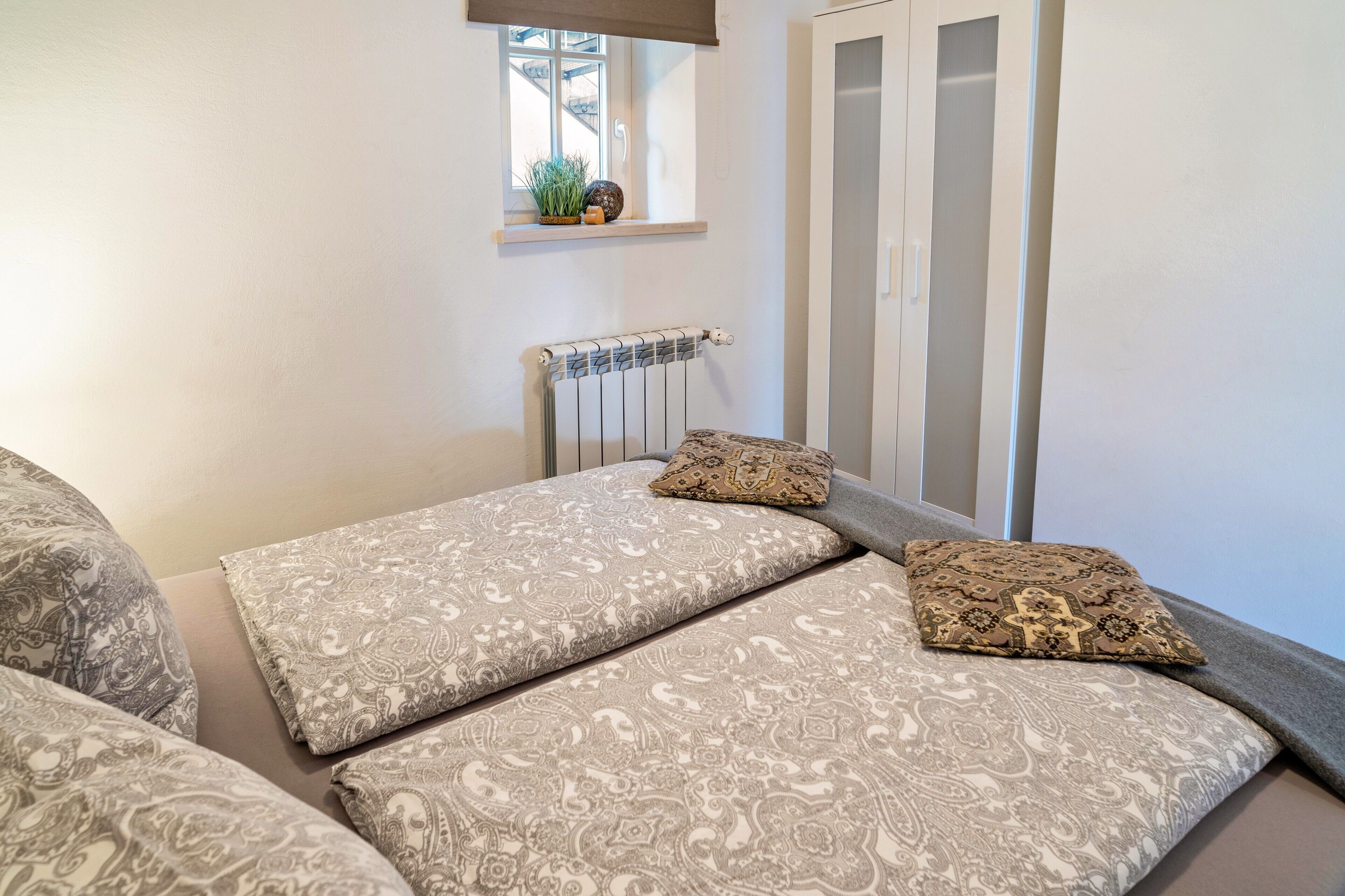 Un dormitorio con cama, almohadas, un radiador, una ventana y un armario.