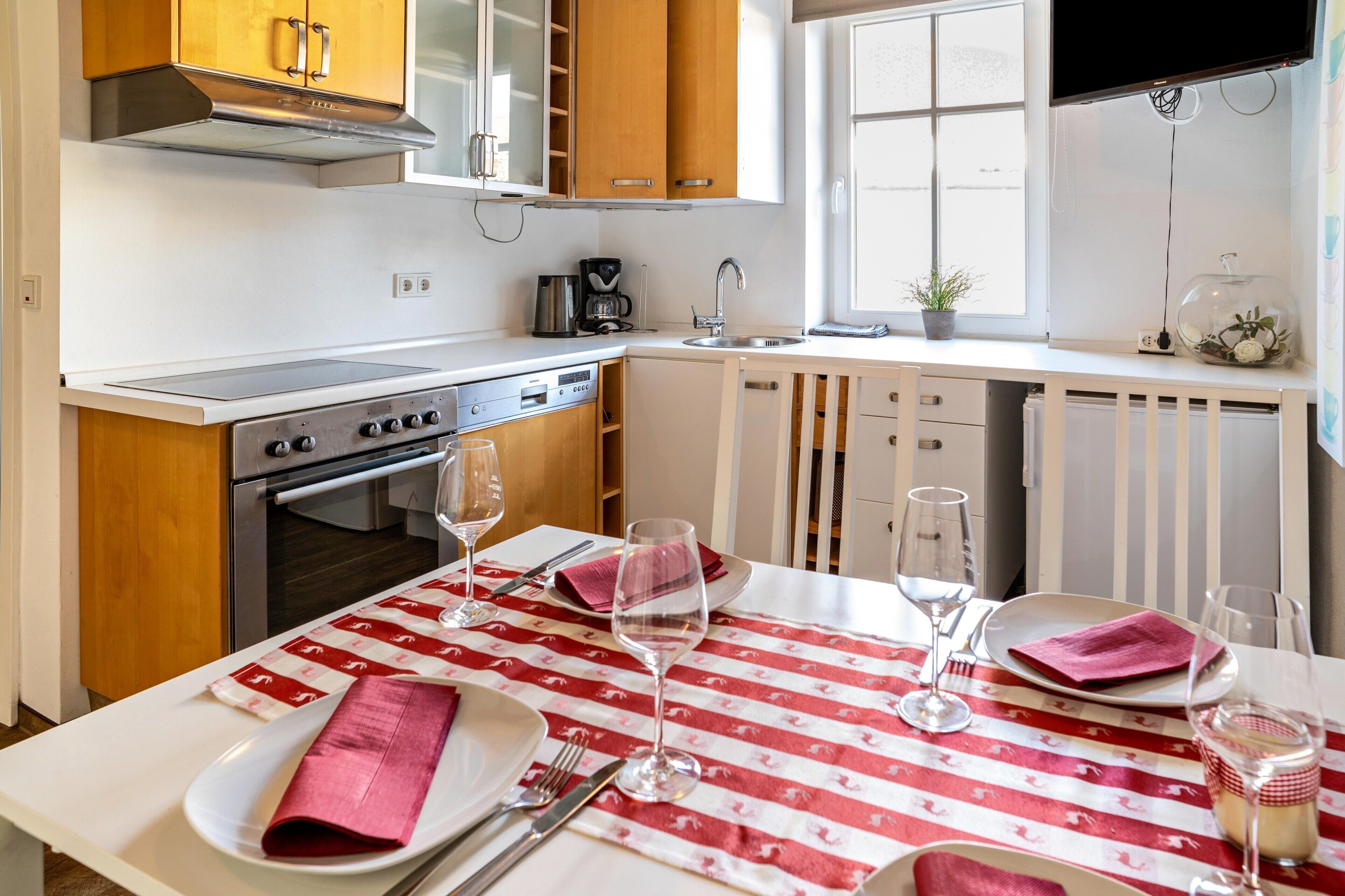 A modern kitchen with a stove, oven, and sink, a dining table set with a red and white tablecloth, and a window with a view.