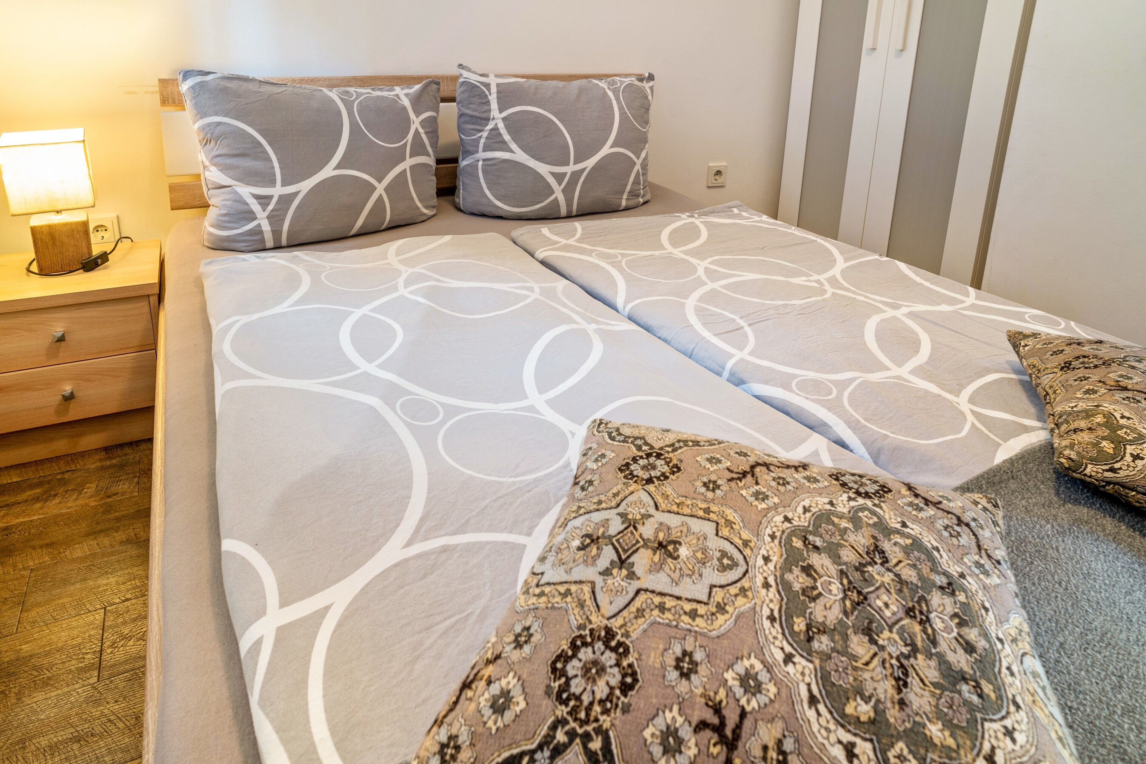 A neatly made bed with patterned bedding, a bedside table with a lamp, and a wooden floor.