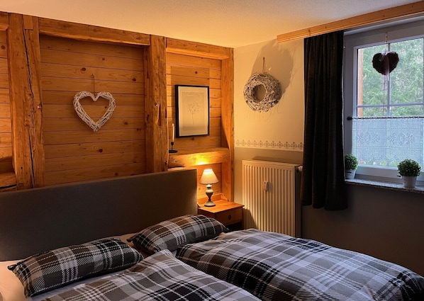 A bedroom with a wooden headboard, a bedside table with a lamp, a framed picture, a heart-shaped decoration, and a window with a view.