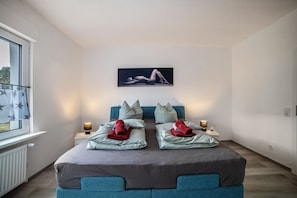 A modern bedroom with a large bed, two bedside tables, a wall-mounted artwork, and a window with curtains.