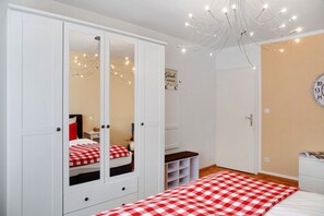 Une chambre avec une grande armoire, un lit recouvert d’une couverture à carreaux, une table de chevet, un miroir, une horloge au mur et une guirlande lumineuse.