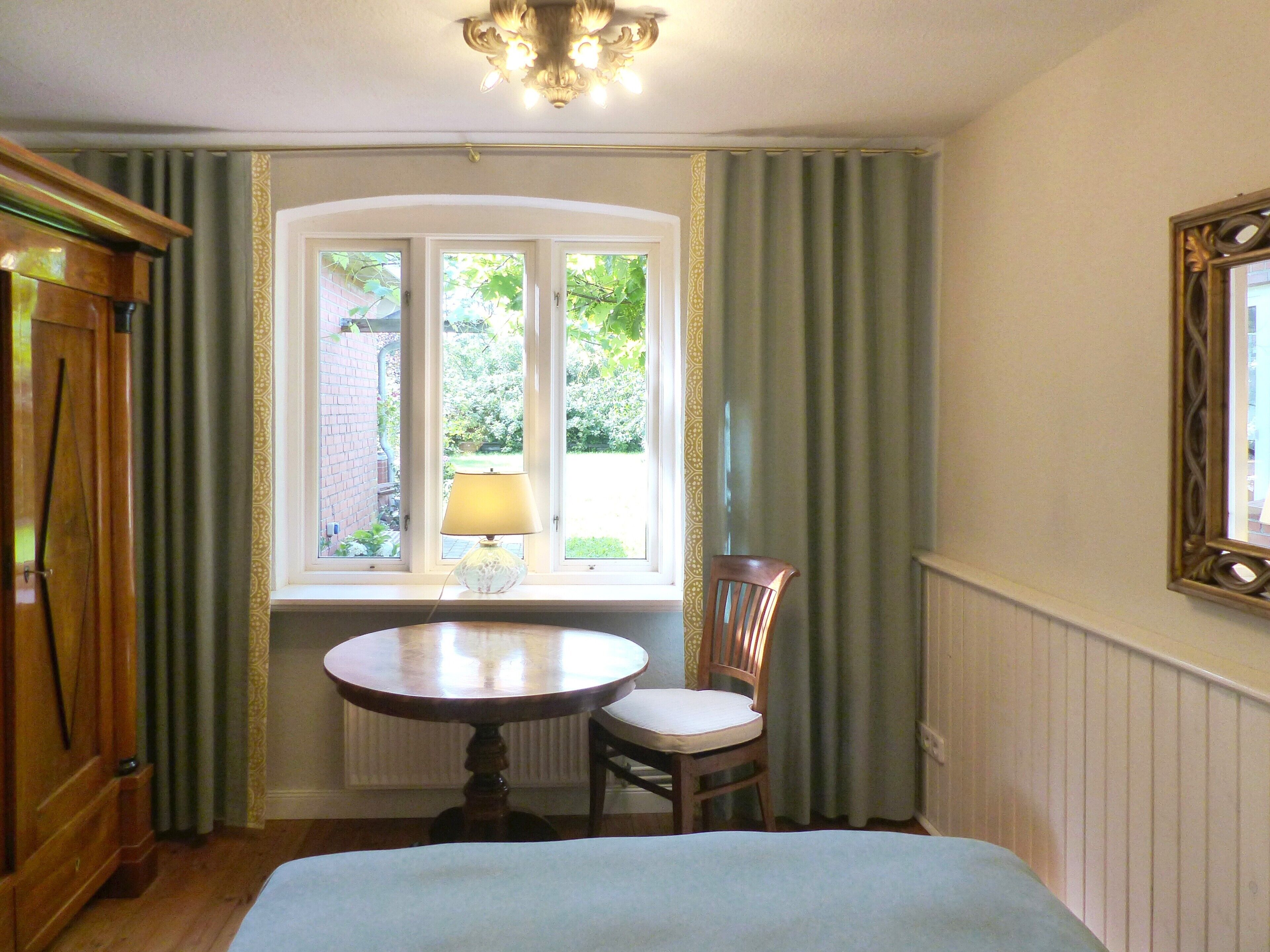 Een kamer met een ronde tafel, een stoel, een raam met gordijnen, een lamp en een spiegel aan de muur.