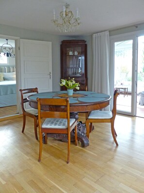 Uma sala de jantar com uma mesa redonda, quatro cadeiras, um lustre e vista através de uma porta de vidro.