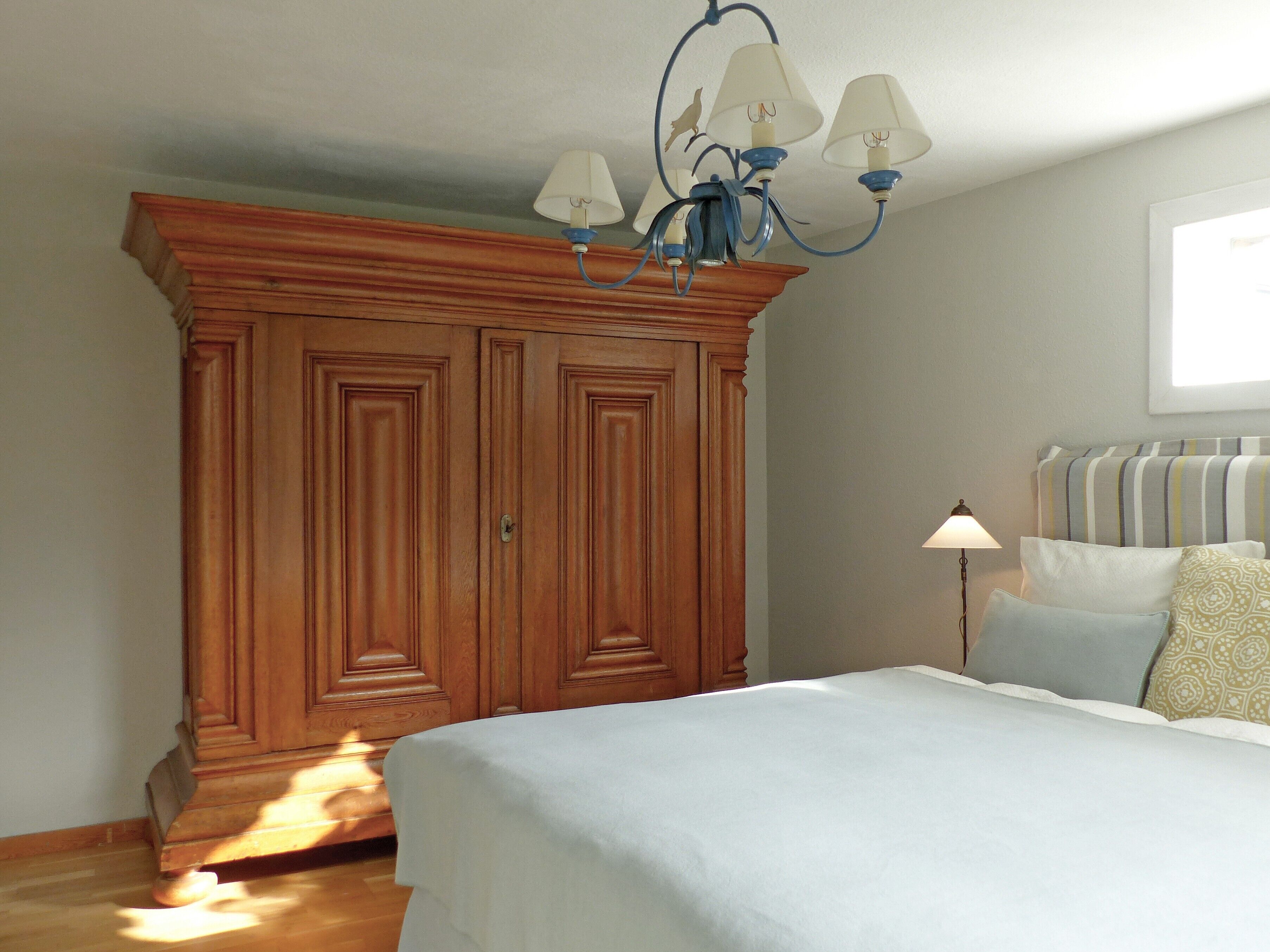 Een slaapkamer met een grote houten garderobe, een bed met een witte dekbedovertrek, een blauwe kroonluchter en een raam met jaloezieën.