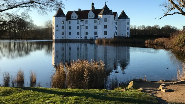 Een groot, wit, kasteelachtig gebouw met meerdere torens, omgeven door een rustig meer en groene velden.