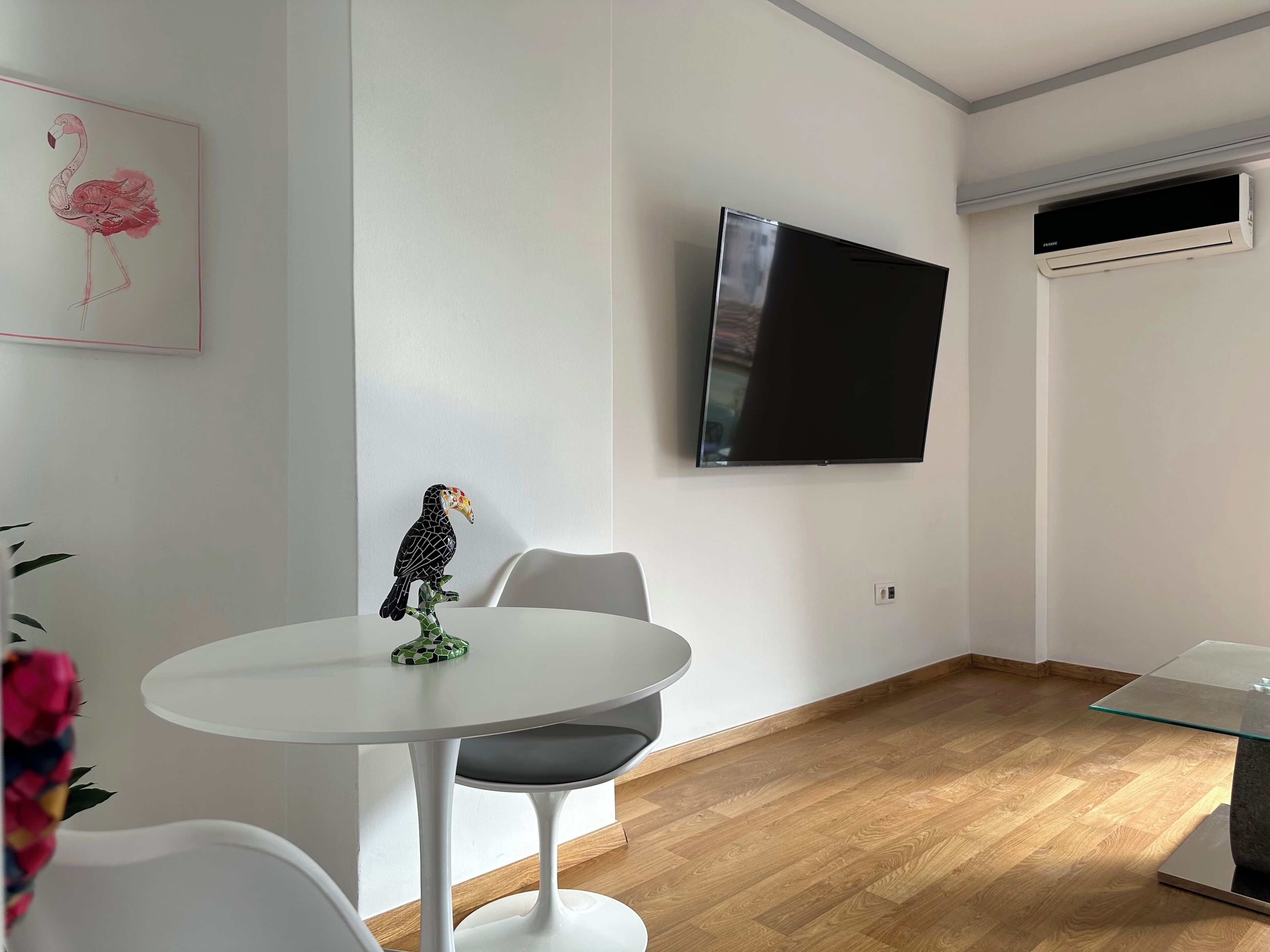 A modern room with a flat-screen TV mounted on the wall, a white dining table with chairs, and a wooden floor.