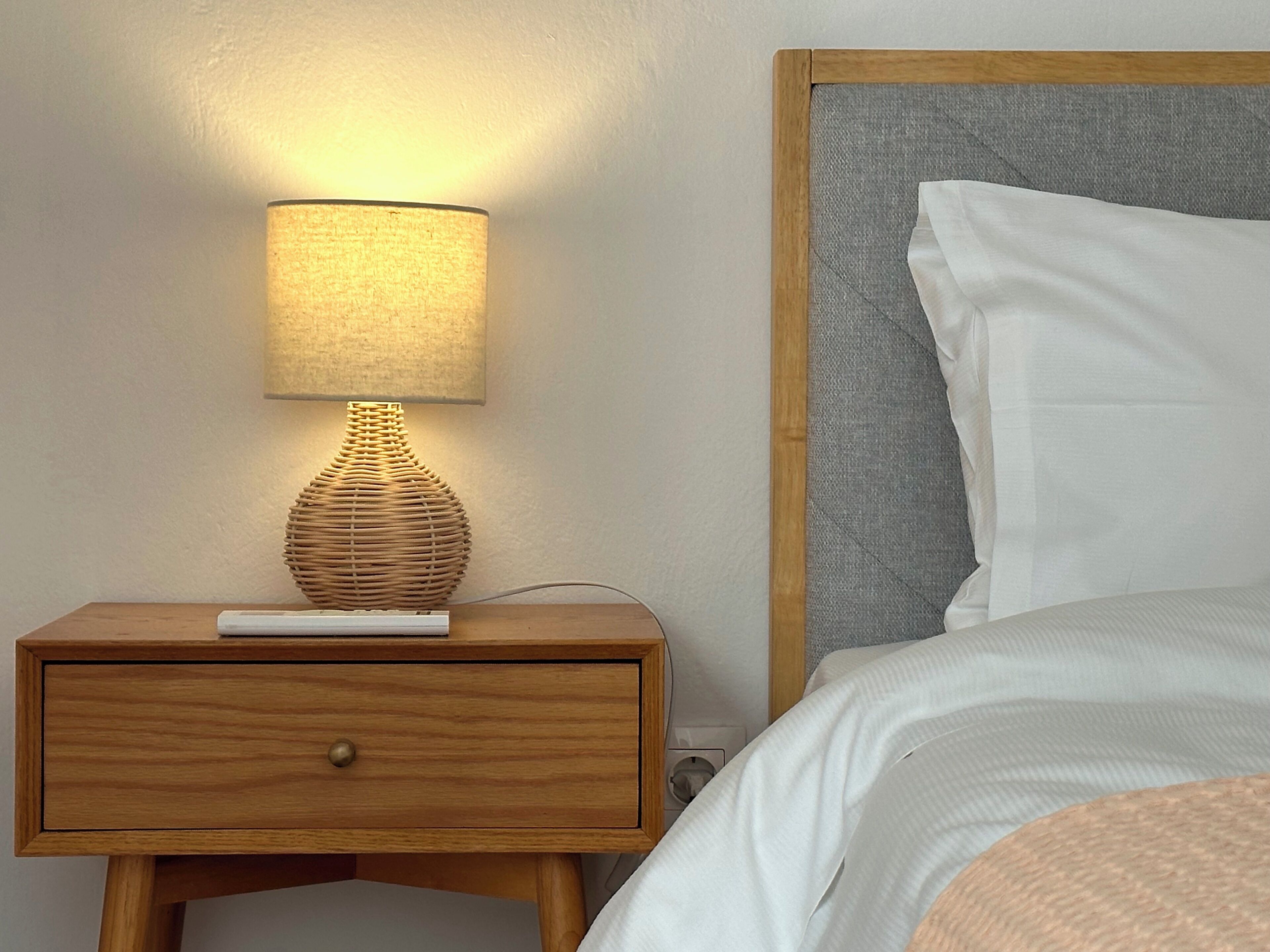 Una mesita de noche de madera con una lámpara, un almohadón blanco y una cama con cabecero gris.
