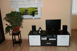 Une pièce avec un téléviseur posé sur un meuble, une petite table avec une plante, une fenêtre donnant sur un paysage verdoyant et un meuble blanc avec des tiroirs.