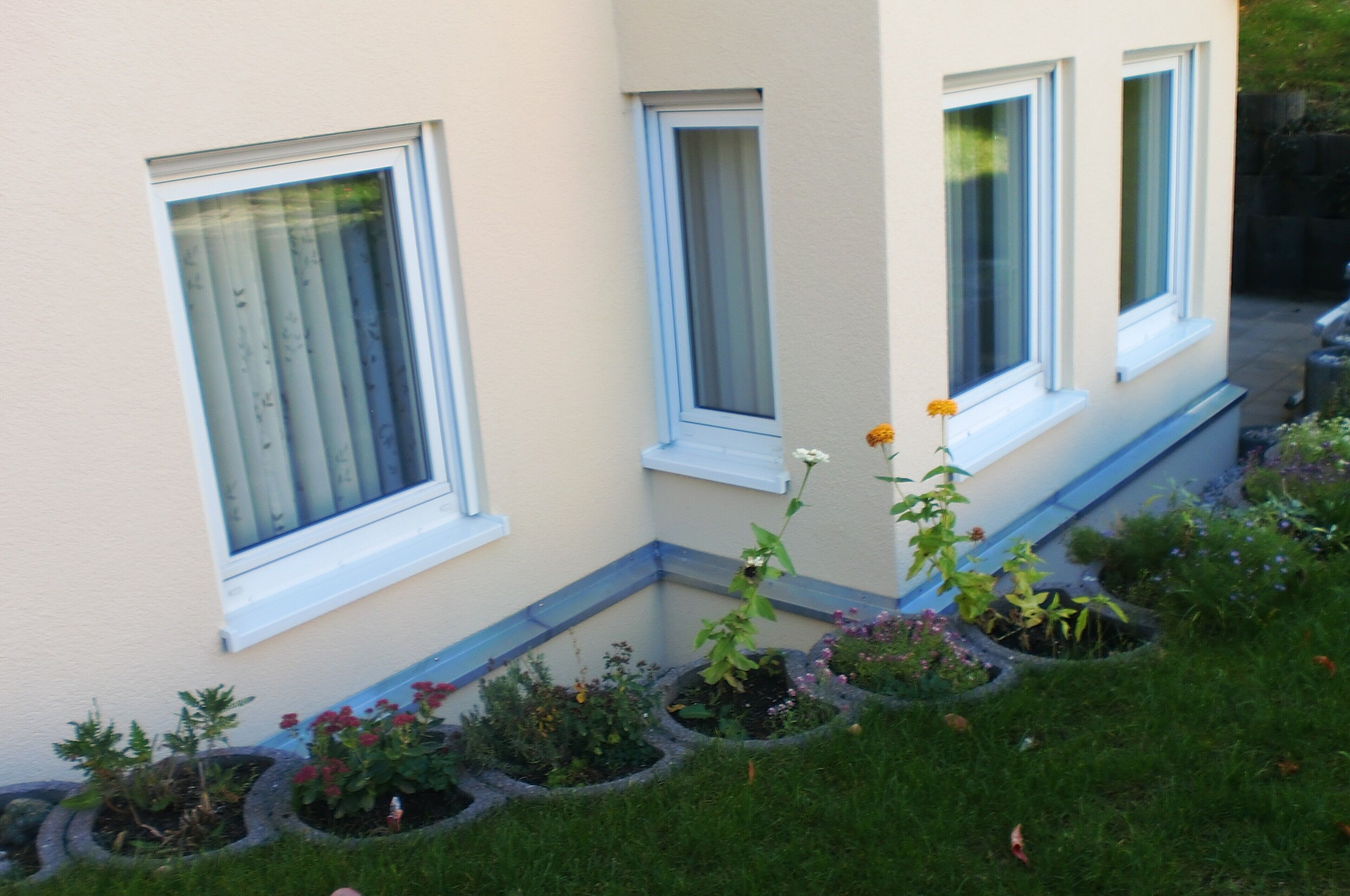 A building with three windows, a small garden with various plants, and a pathway leading to the entrance.