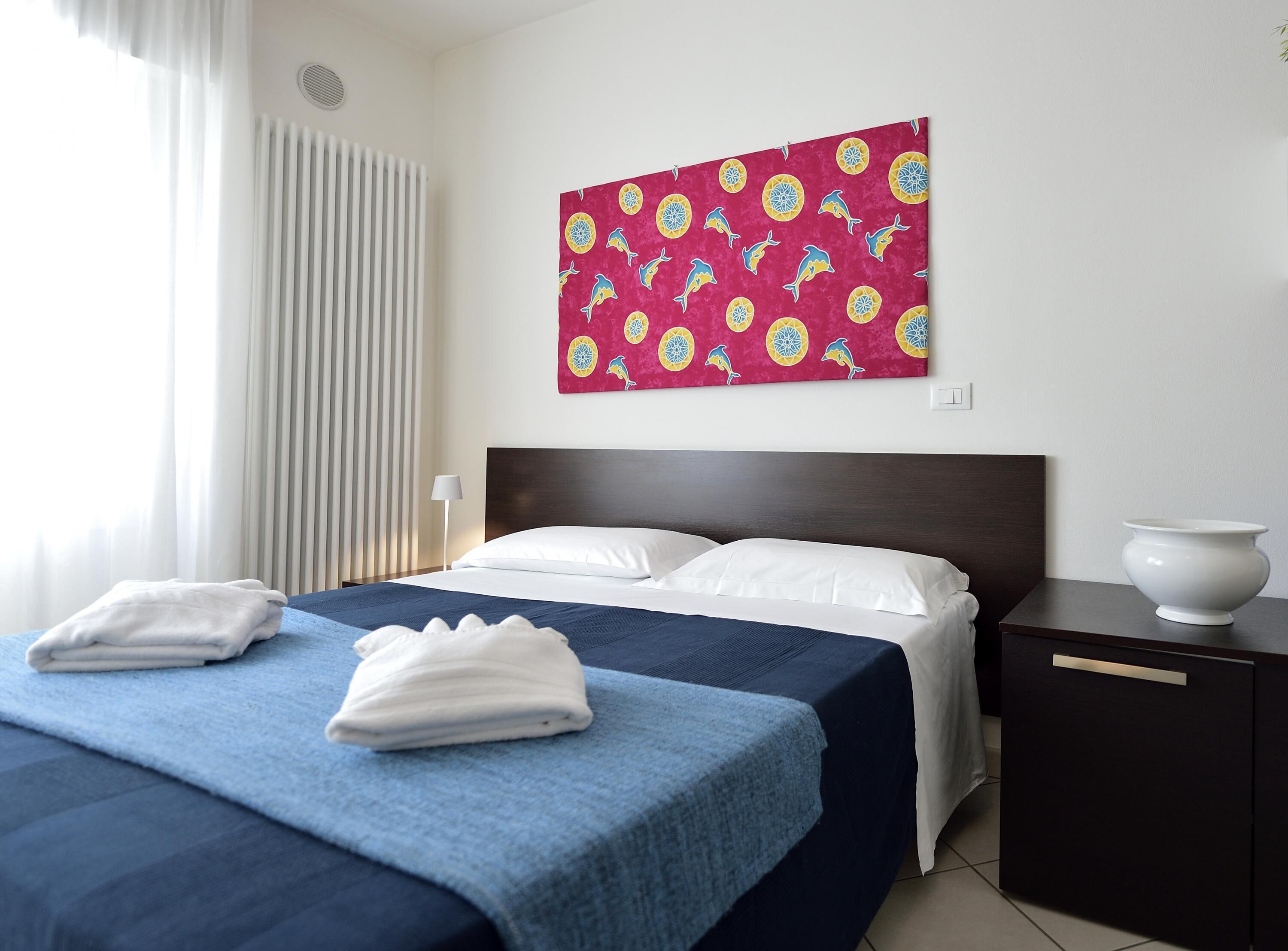 Une chambre d’hôtel avec un lit, des oreillers blancs, une couverture bleue, une serviette blanche, une table de chevet et un vase blanc.