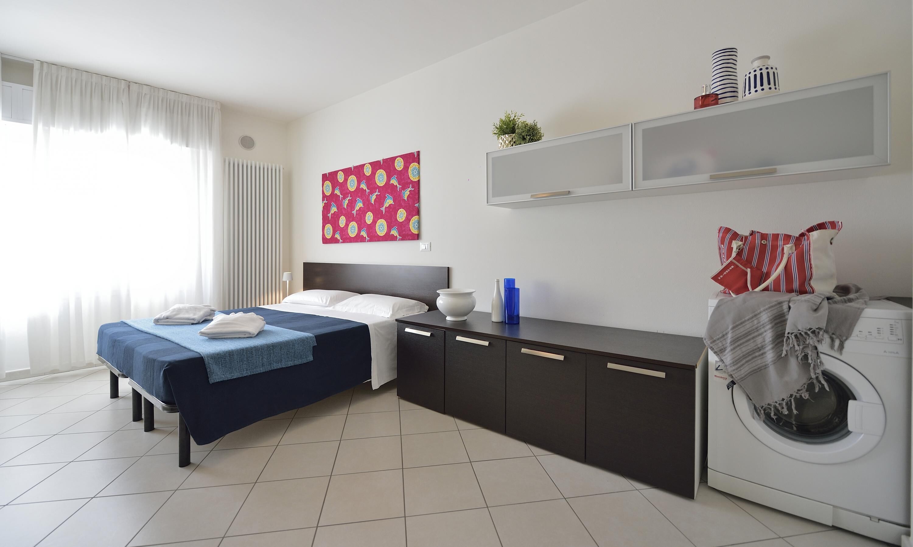 A hotel room with a bed, a washing machine, a cabinet, and a shelf with decorative items.