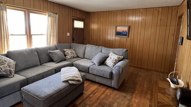 Een woonkamer met een hoekbank, een klein poufje, houten vloeren en een wand bekleed met hout.