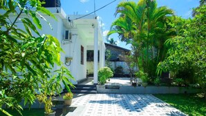 Un patio con sendero adoquinado, un edificio blanco y exuberante vegetación, incluyendo palmeras.