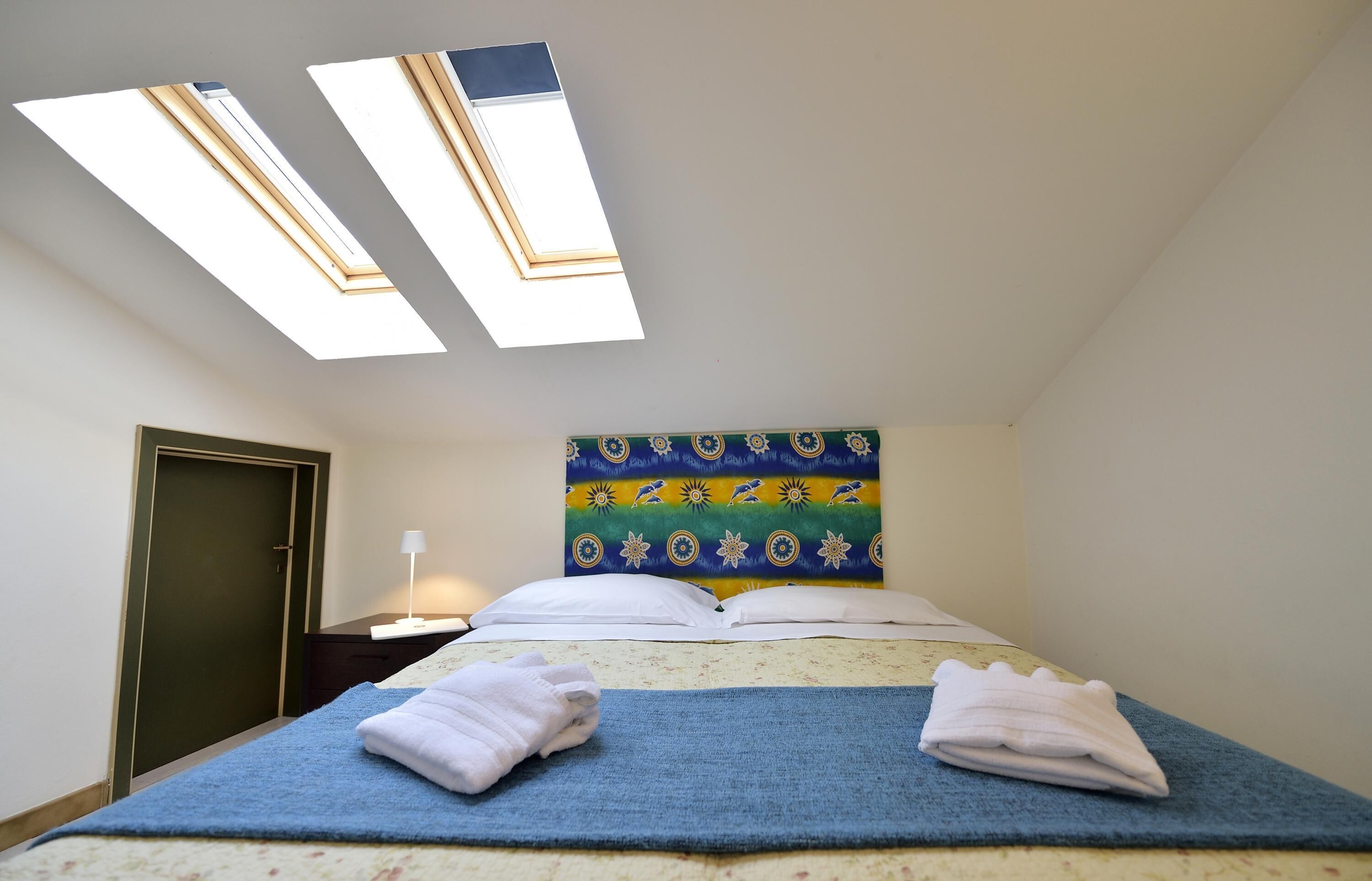 A bedroom with a bed, two skylights, a nightstand, a lamp, and a door.