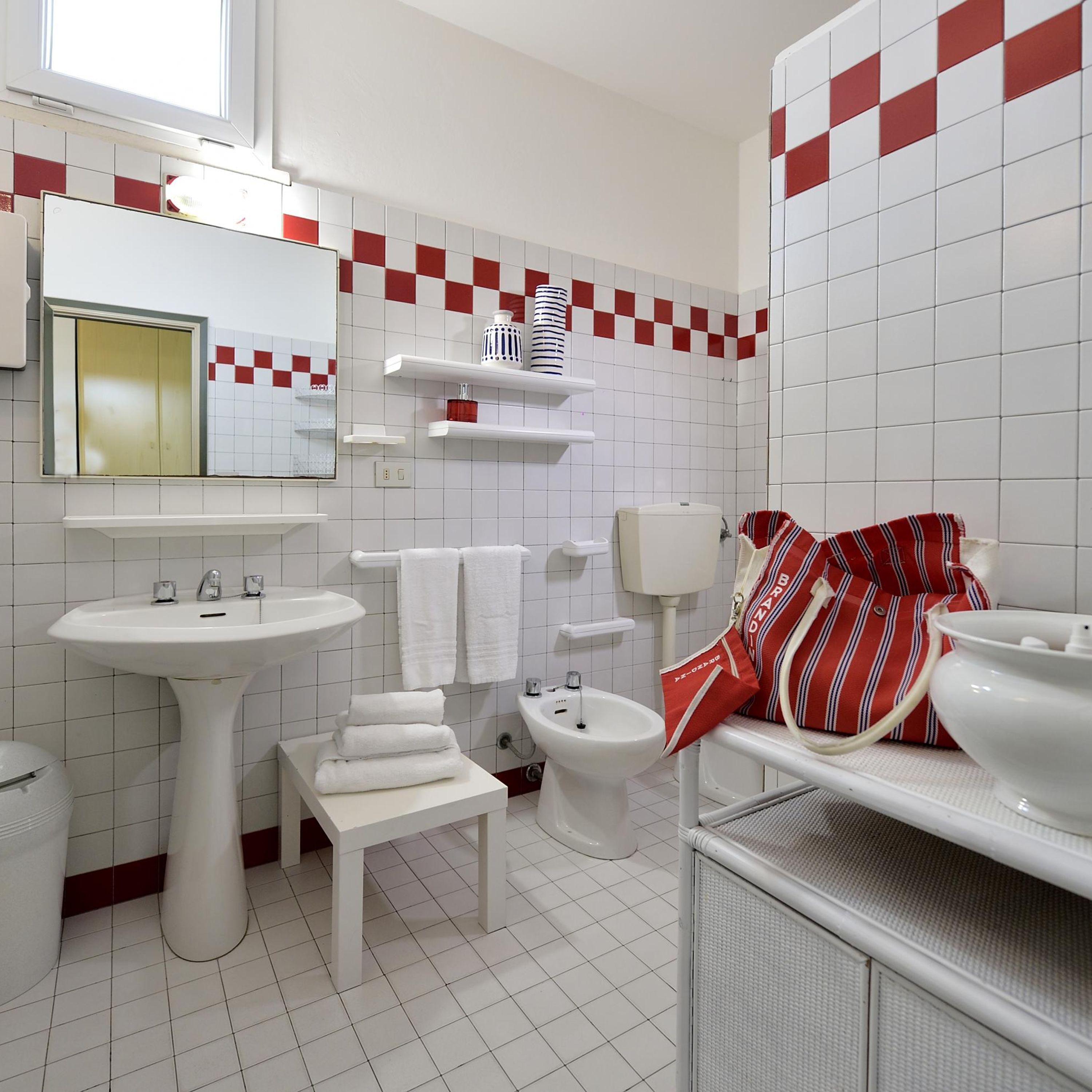 Ein Badezimmer mit weißen Fliesen, einem Waschbecken, einer Toilette und einem Bidet. Auf der Ablage liegen Handtücher und ein gestreiftes Tuch.