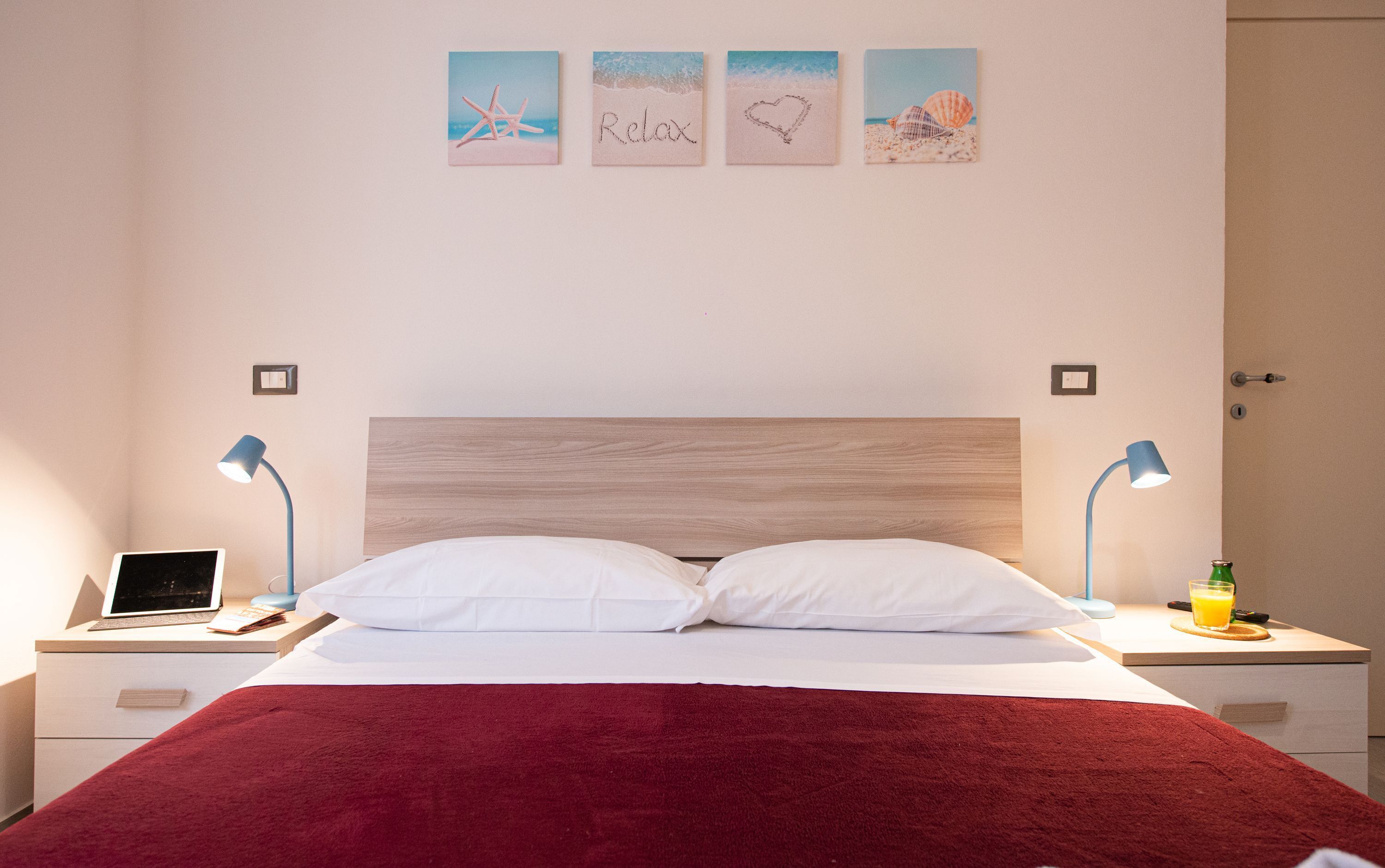 A bedroom with a wooden headboard, two bedside tables with lamps, a laptop, a glass of orange juice, and a red blanket.
